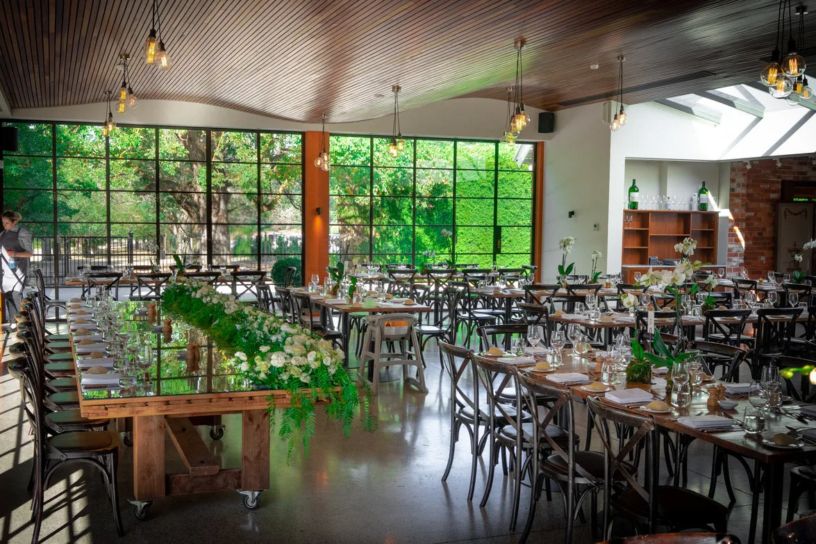 Wide venue with long tables, greenery runners, and a dramatic wavy timber ceiling