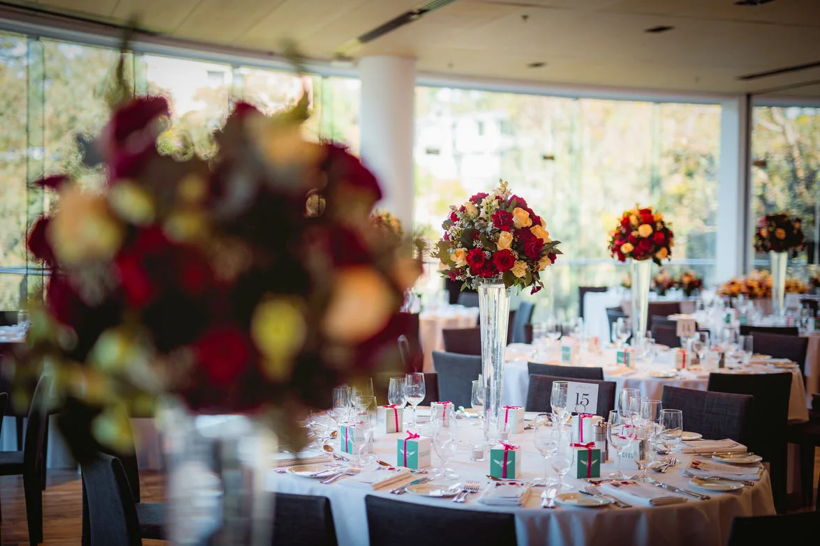 Sunlit glass-walled venue with soft-focus red bloom and tall centrepieces