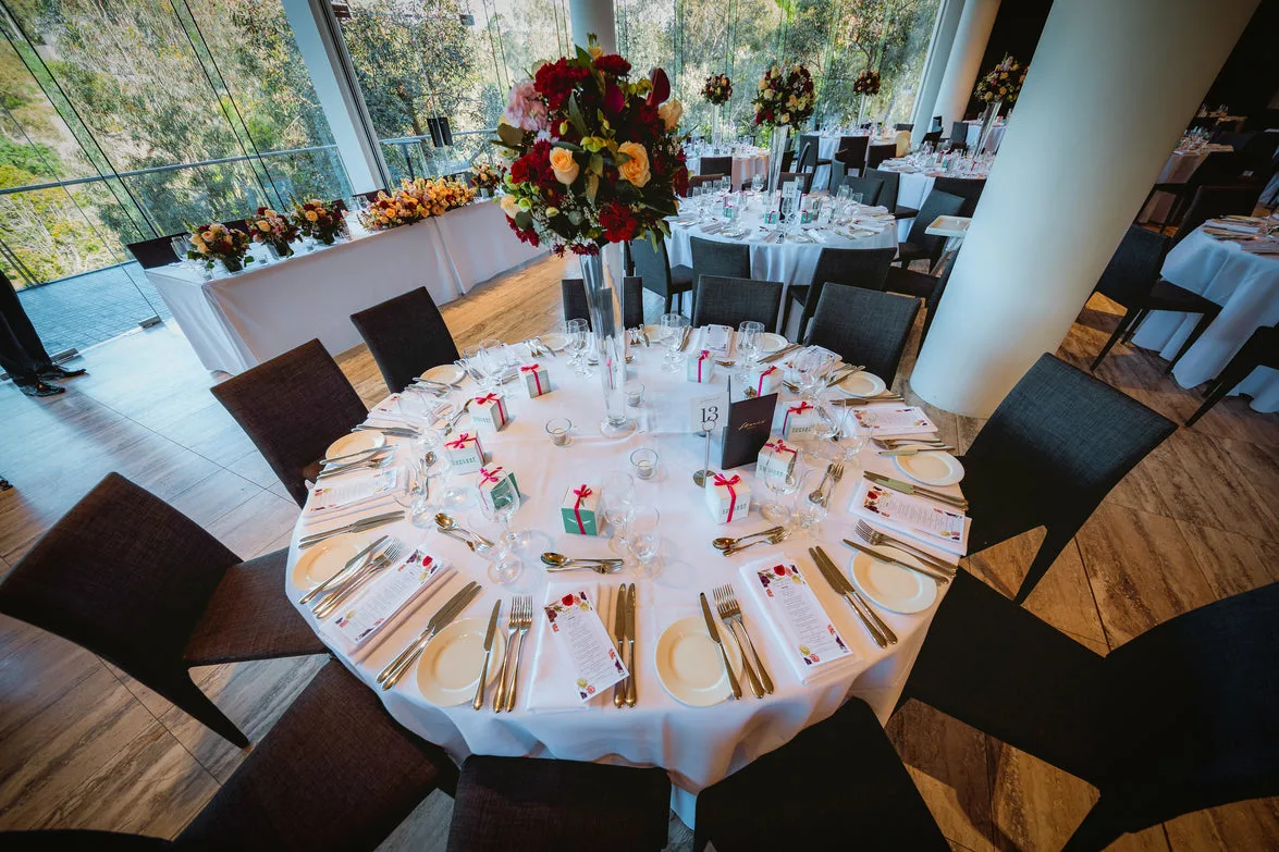 Bright reception hall with round tables, red and peach centrepieces, and bush views